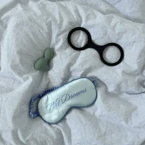 Billede af en dyne. På dynen ligger sorte silikone håndjern, en grøn finger vibrator og lyseblå natbriller med skriften "Wet Dreams".